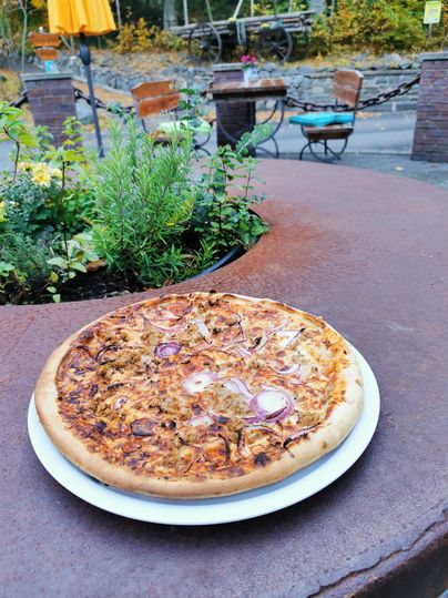 Eine frische Pizza auf einem rustikalen Tisch. Im Hintergrund sind einige Sitzgelegenheiten und Pflanzen sichtbar.