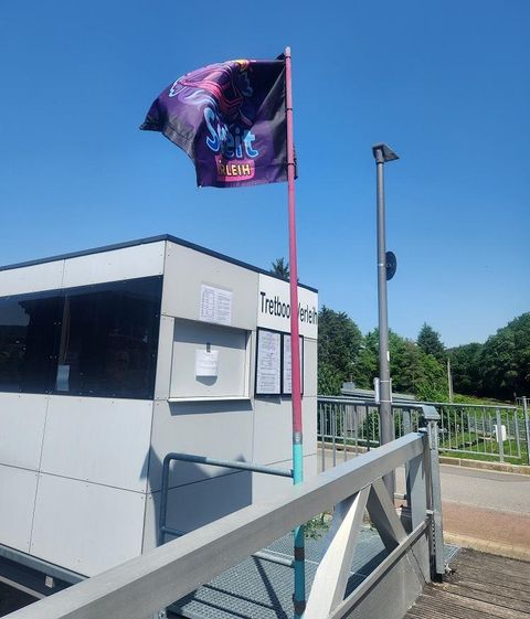 Un kiosque au bord de l'eau avec un drapeau coloré flotte dans le vent. En arrière-plan, on voit des arbres et un ciel bleu.