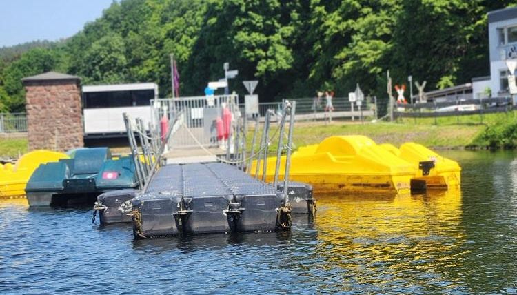 Un plan d'eau rempli de bateaux avec un ponton au premier plan. Sur la rive, il y a des canoës jaunes et des barques vertes.