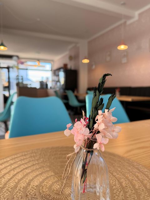 Ein gemütliches Café mit blauen Stühlen und warmer Beleuchtung. Auf dem Tisch steht eine kleine Glasvase mit pinken Blumen.