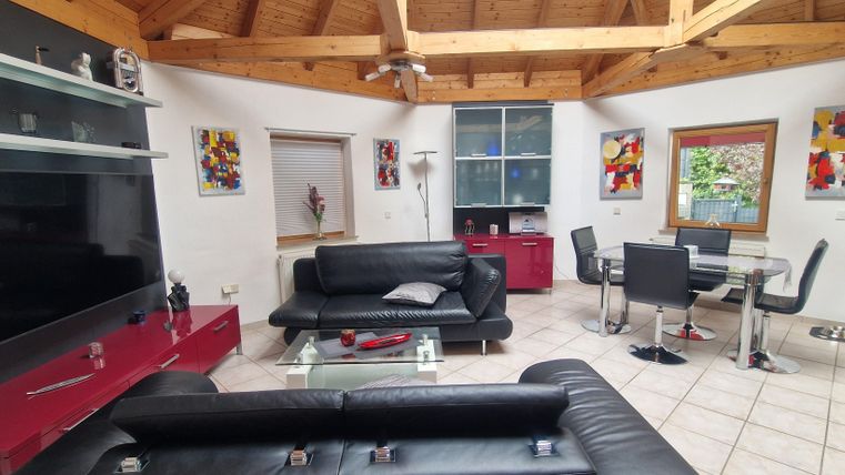 Ein modern gestaltetes Wohnzimmer mit schwarzen Sofas und einem großen Fernseher. Der Raum hat Holzdecken und bunte Kunstwerke an den Wänden.