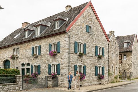 Ein charmantes Steinhaus mit Fensterläden in Blau und blühenden Pflanzen. Zwei Personen stehen auf dem Gehweg und unterhalten sich.