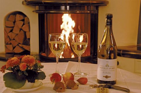 Ein gemütlicher Tisch mit zwei Weingläsern, einer Flasche Sekt und frischen Äpfeln. Im Hintergrund brennt ein Kamin und sorgt für eine warme Atmosphäre.