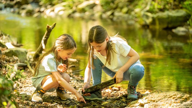 Deux filles jouent au bord d'un petit ruisseau. Elles examinent ensemble un bâton dans l'eau.