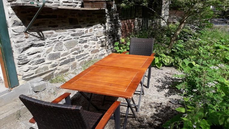 Une table en bois avec deux chaises se trouve dans un espace extérieur verdoyant. En arrière-plan, un mur en pierre et des plantes sont visibles.