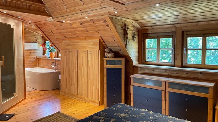 Een gezellige slaapkamer met houten plafond en grote vensters. Op de achtergrond is een moderne badkamer met een badkuip te zien.