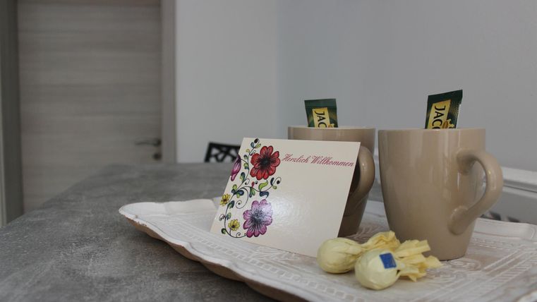 Zwei Tassen auf einem Tablett, begleitet von einer Willkommenskarte und kleinen Süßigkeiten. Im Hintergrund ist ein heller Raum zu sehen.