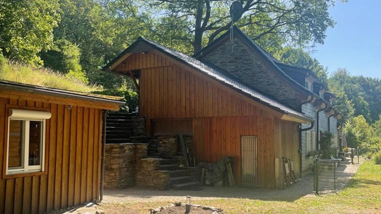 Een rustiek houten huis met een overdekt dak naast een ander gebouw. De tuin is groen en omgeven door bomen.