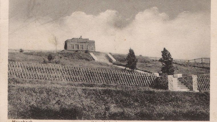 Un monument aux morts situé sur une colline, avec un petit bâtiment. Au premier plan, on aperçoit des escaliers et une clôture en tôle ondulée.