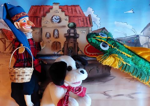 Eine bunte Puppenspiel-Szene mit einer Marionette, einem Stoffhund und einem grünen Drachen. Im Hintergrund ist ein malerischer Brunnen und ein Gebäude zu sehen.