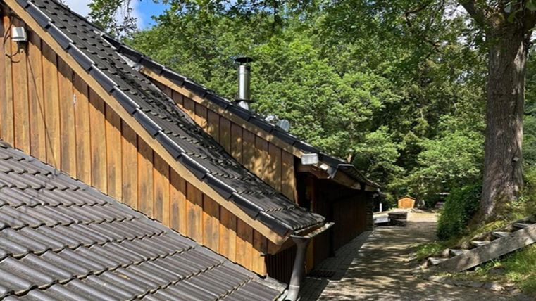 Een rustiek huis met een schuin, betegeld dak en houten bekleding. Op de achtergrond zijn bomen en een smal pad te zien.