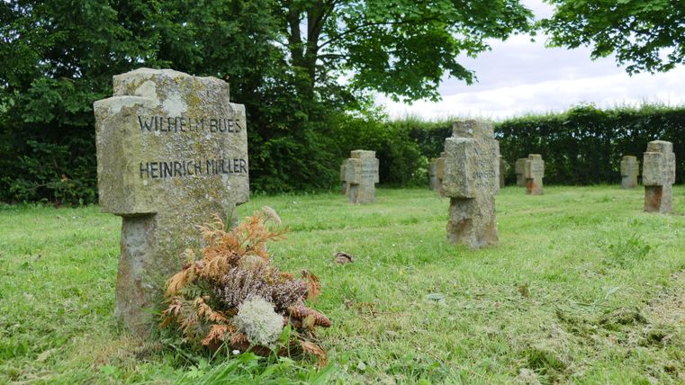 Un cimetière avec plusieurs pierres tombales en pierre, situé dans un pré verdoyant. Certaines pierres tombales portent des noms gravés et un petit bouquet de fleurs est déposé sur l'une des tombes.