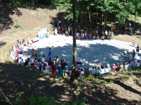 Eine Gruppe von Menschen versammelt sich in einem Wald für eine Veranstaltung. Sie stehen an einem runden Platz, umgeben von Bäumen.
