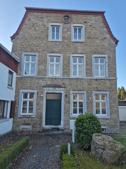 Une maison historique en pierre avec une porte d'entrée verte et de grandes fenêtres. Entourée d'un petit jardin et de quelques pierres au premier plan.