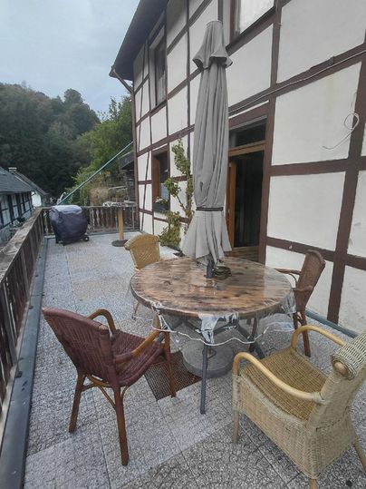 Une terrasse avec une table en bois ronde et plusieurs chaises. En arrière-plan, on peut voir une maison à colombages traditionnelle.
