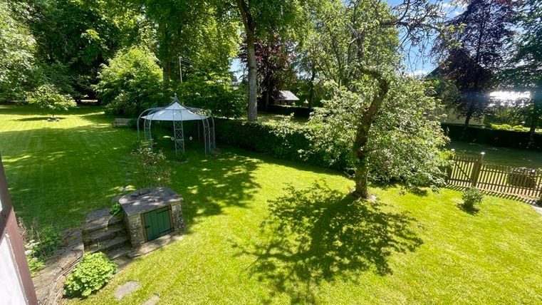 Ein gepflegter Garten mit grünem Gras, einem Pavillon und einem großen Baum. Im Hintergrund sind weitere Bäume und eine eingezäunte Fläche sichtbar.
