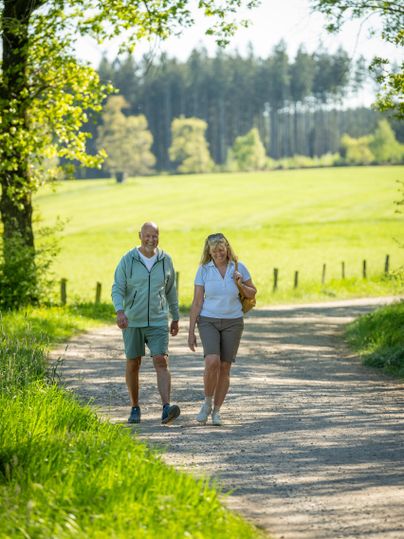 Een ouder echtpaar wandelt op een rustige weg door een groene omgeving. De zon schijnt en de bomen bieden schaduw.