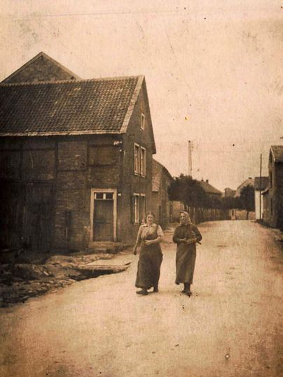 Deux femmes marchent dans une rue calme, bordée de bâtiments anciens. Le décor dégage une atmosphère nostalgique et semble historique.