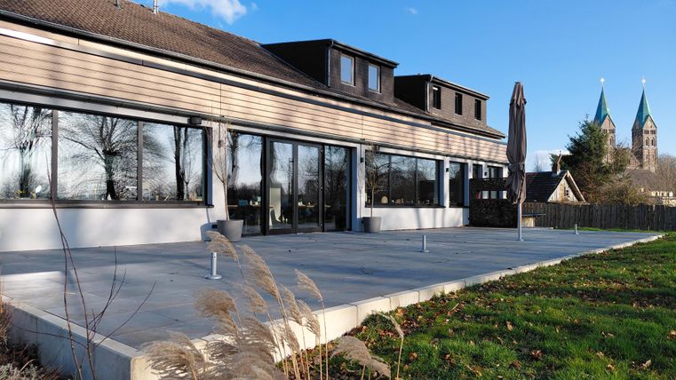 Ein modernes Gebäude mit großen Fenstern und einer Terrasse. Im Hintergrund sind zwei Kirchtürme und ein klarer blauer Himmel zu sehen.
