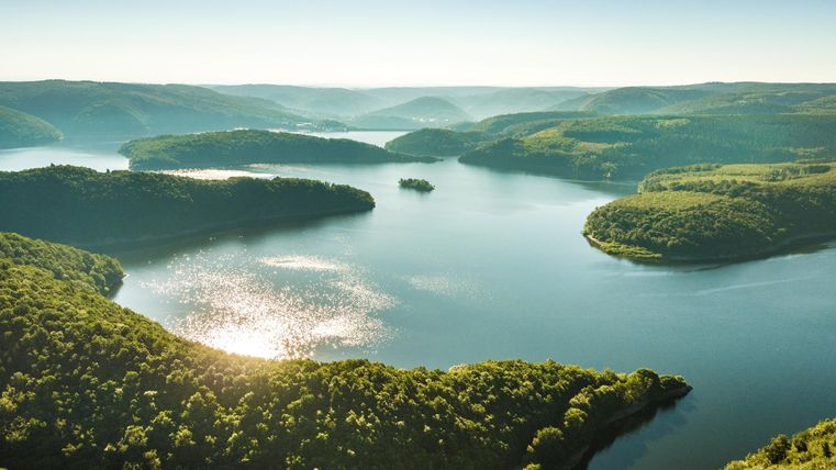 Eine malerische Landschaft mit einem ruhigen See und sanften Hügeln. Die Bäume reflektieren das Licht der Sonne und schaffen eine friedliche Atmosphäre.