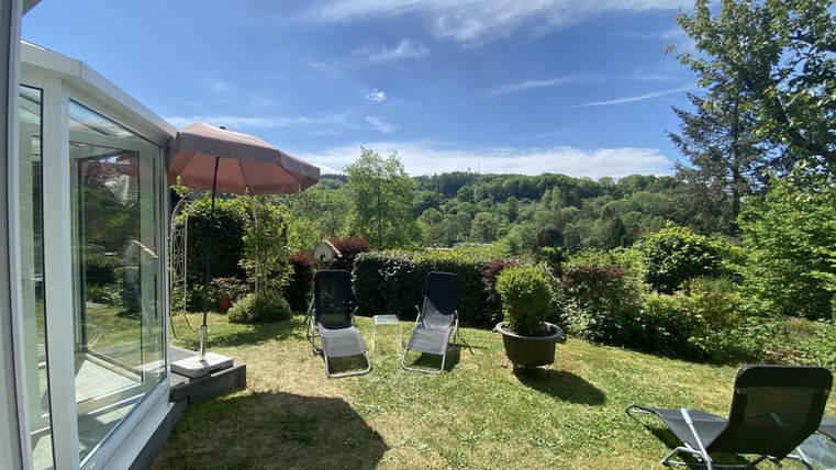 Een mooie tuin met ligstoelen en een parasol. Op de achtergrond zijn heuvels en bomen zichtbaar onder een heldere blauwe lucht.