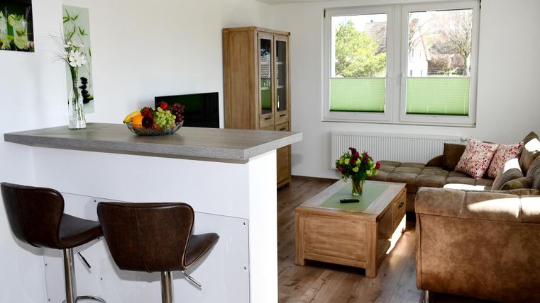 Une cuisine-salon lumineuse et moderne avec un canapé confortable et une table basse. À l'arrière-plan, on voit de grandes fenêtres qui laissent entrer beaucoup de lumière naturelle.