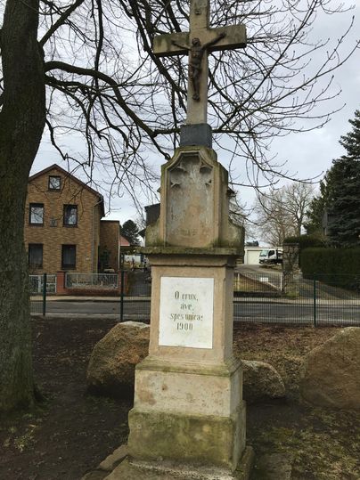 A historic cross stands on a stone pedestal. Several buildings and trees are visible in the background.