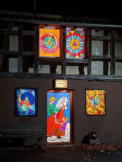 Bunte, beleuchtete Fenster und eine farbenfrohe Tür an einem Gebäude. Die Fenster zeigen verschiedene geometrische und figurative Motive.