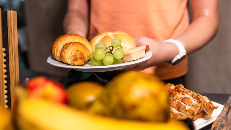 Eine Person hält einen Teller mit frischen Brötchen, Trauben und einem Croissant. Im Hintergrund sind verschiedene Früchte sichtbar.