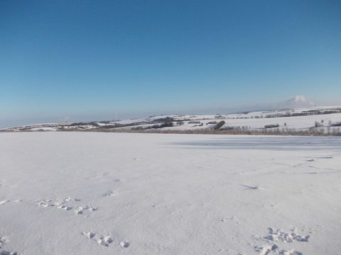 Eine weite, schneebedeckte Landschaft unter einem klaren blauen Himmel. Im Hintergrund sind sanfte Hügel sichtbar.