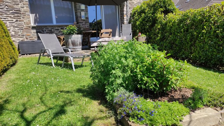 Ein Garten mit einer schönen Sitzecke und Sonnenschirm. Im Vordergrund wachsen verschiedene Pflanzen und Kräuter.