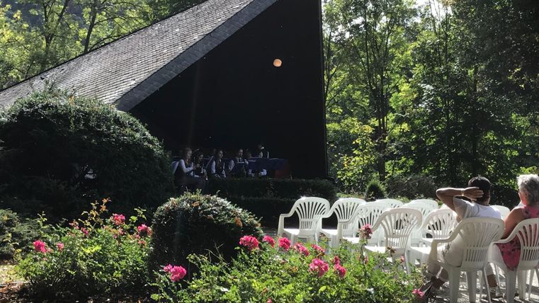 Eine Bühne im Freien, umgeben von Bäumen und Blumen. Im Hintergrund sitzen Menschen in weißen Stühlen.