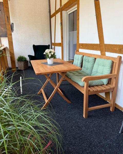 Een gezellige zithoek op een balkon met een houten tafel en een bank. Op de tafel staat een kleine vaas met bloemen en op de achtergrond zijn planten zichtbaar.