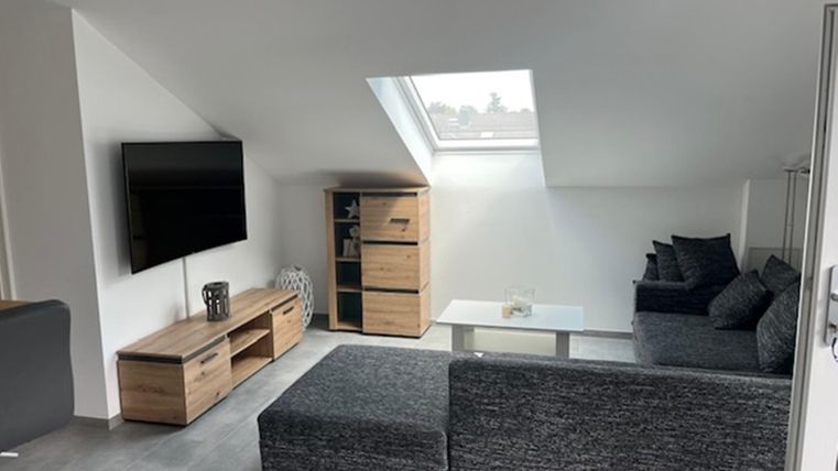 Ein modernes Wohnzimmer mit einem gemütlichen Sofa und einem großen Fernseher. Das Zimmer hat helle Wände und viel natürliches Licht durch das Dachfenster.