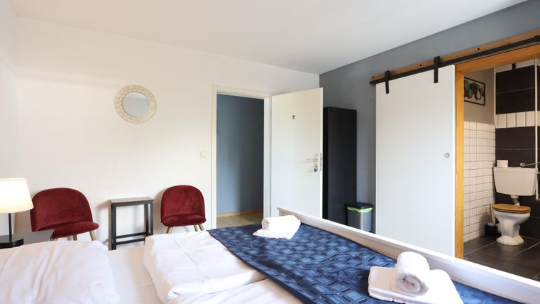 Ein modernes Schlafzimmer mit einem Doppelbett und zwei roten Stühlen. Im Hintergrund sind ein Badezimmer und eine große Schiebetür zu sehen.