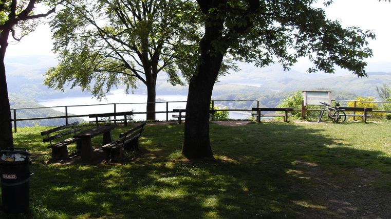 Eine ruhige Aussicht mit Bäumen und einer Bankgruppe. Im Hintergrund sieht man eine Landschaft mit Wasser und Hügeln.