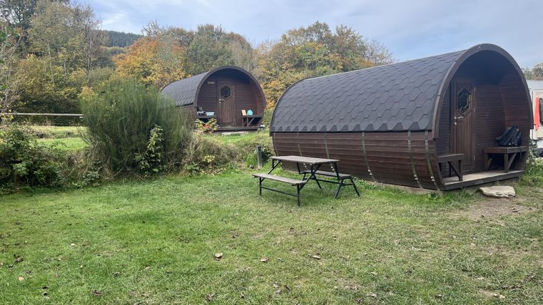 Zwei gemütliche Hütten stehen in einer grünen Wiese, umgeben von Bäumen. Das Wetter ist bewölkt, und es gibt eine kleine Sitzecke im Freien.
