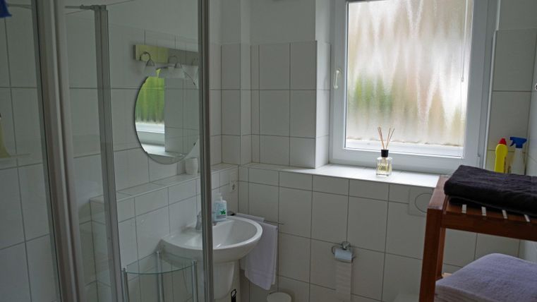 Ein modernes Badezimmer mit weißen Fliesen und einer Dusche. Das Fenster sorgt für Tageslicht und mehrere Pflegeprodukte sind sichtbar.
