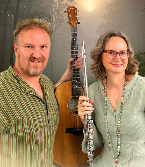 Ein Mann mit einer Gitarre und eine Frau mit einer Flöte stehen zusammen vor einem malerischen Waldhintergrund. Beide lächeln freundlich und tragen legere Kleidung.