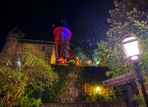 Eine alte Burg erleuchtet von bunten Lichtern bei Nacht. Im Vordergrund ist eine Straßenlampe und ein Schild mit dem Namen "Moritz-Kraus-Platz".