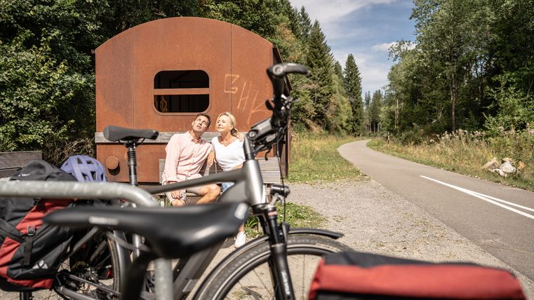 Ein Paar sitzt auf einer Bank neben einem rostigen Eisenbahnwagen, mit Fahrrädern im Vordergrund und einem Waldweg im Hintergrund.