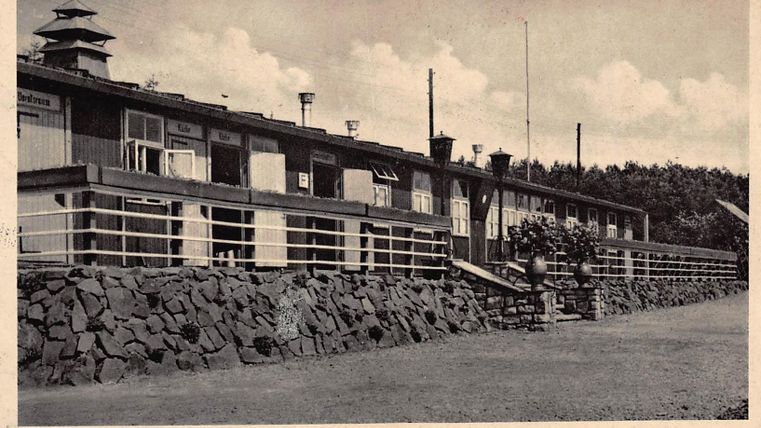 Ein historisches Gebäude mit mehreren Fenstern und einer Veranda. Umgeben von einem gepflegten Gelände und Bäumen im Hintergrund.