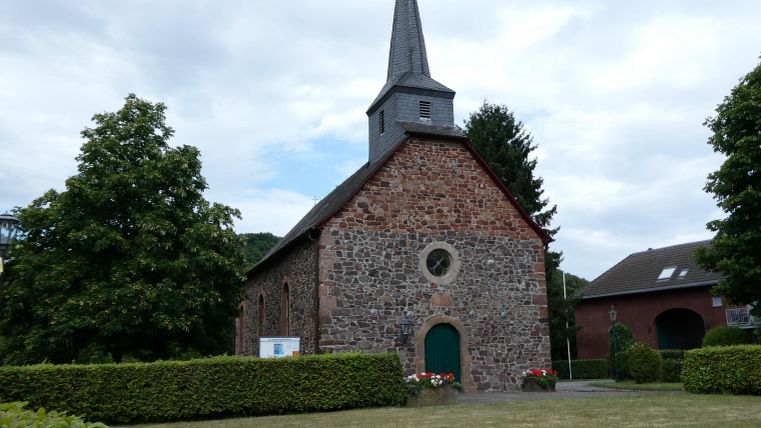 A small church with a pointed tower and a surrounding garden. The ground is landscaped with grass and bushes.