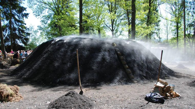 Ein Holzkohleofen in einem Waldgebiet, umgeben von Bäumen. Der Ofen ist mit Holz und Erde abgedeckt, während Dampf aufsteigt.