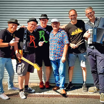 Zes muzikanten staan glimlachend voor een heldere, geribbelde muur. Ze houden muziekinstrumenten vast en dragen verschillende, kleurrijke kleding.