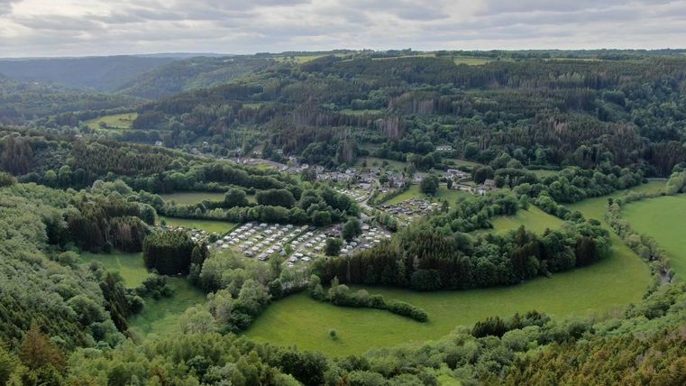 Eine beeindruckende Landschaft mit grünen Hügeln und einem kleinen Dorf in der Talsohle. Im Vordergrund sind Campingplätze und Wiesen zu sehen, umgeben von dichtem Wald.