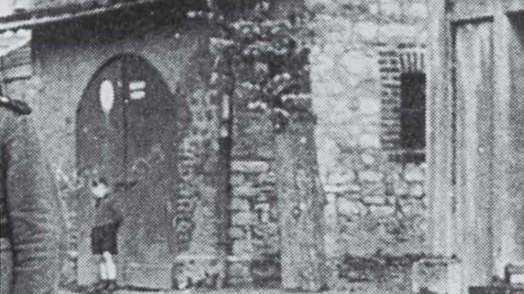 Un garçon se tient devant une vieille porte encastrée dans un mur de pierre. On aperçoit d'autres bâtiments en arrière-plan.