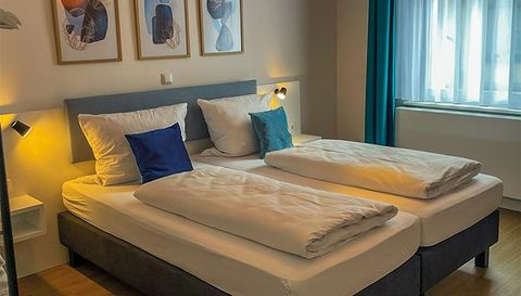 Ein gemütliches Schlafzimmer mit zwei Betten und hellen Kissen. An der Wand hängen dekorative Bilder, und das natürliche Licht strömt durch das Fenster.