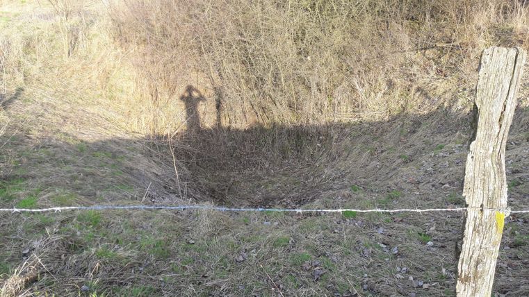 Ein kleiner Graben mit einer unebenen Oberfläche, umgeben von trockenem Gras und Gebüsch. Im Hintergrund ist der Schatten einer Person zu sehen.