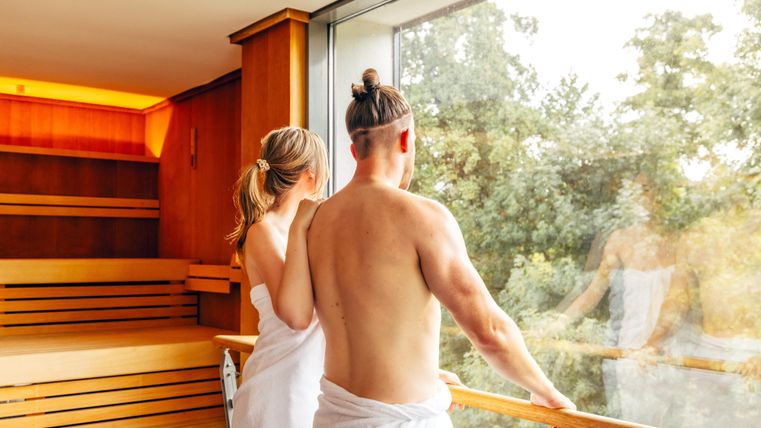 Ein Paar in Saunatüchern steht am Fenster und schaut nach draußen. Die entspannte Atmosphäre wird durch das Holzinterieur und die grüne Natur im Hintergrund betont.